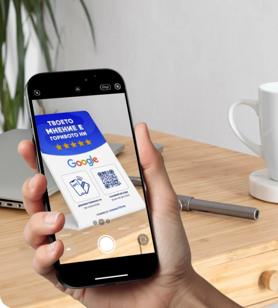 Ръка държи телефон, с който се снима карта за анкета на Google с QR код, поставена върху дървено бюро до чаша и химикалка.
